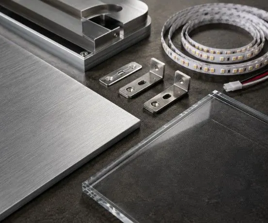 Close-up of brushed aluminium panels, clear acrylic sheets, LED strip lighting, and steel mounting brackets neatly arranged on a workbench.
